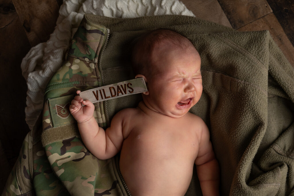 Newborn crying while holding the nametag from her fathers military uniform from Minot AFB, which she is laying on top of.