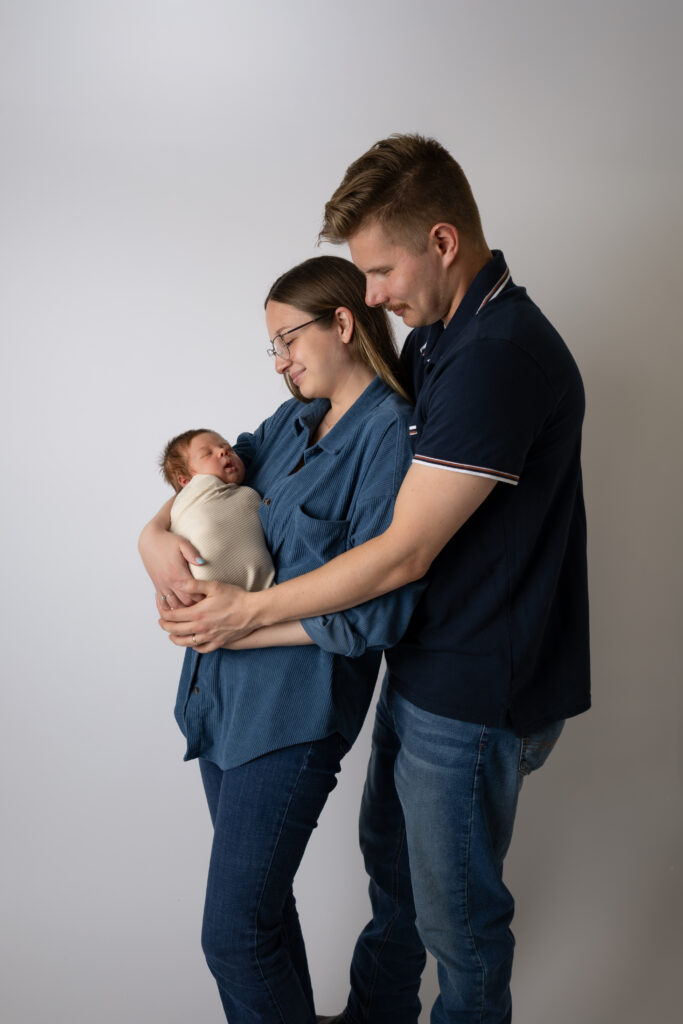 Man with his hands around woman. The woman is holding a newborn in a cream wrap.